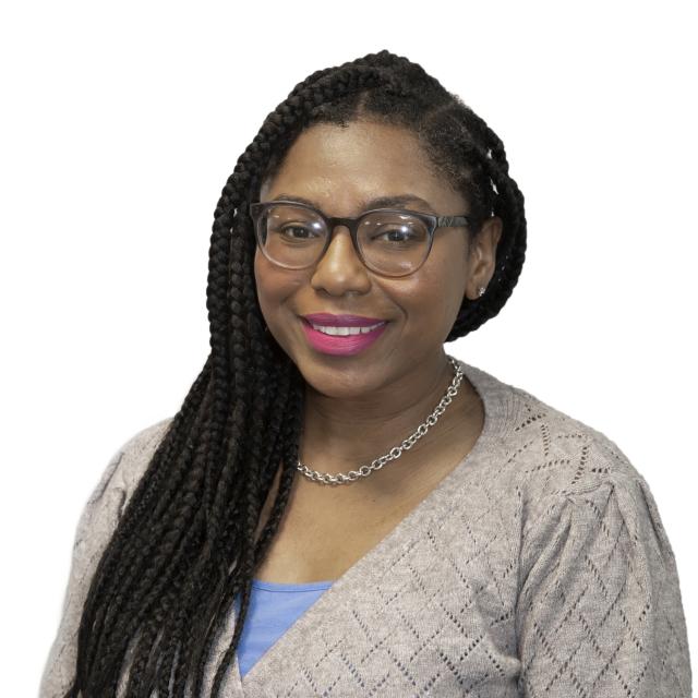 Tiffany Winbush wears an oatmeal cardigan, periwinkle blouse and gold chain as she smiles. She has rounded square glasses on and pink lipstick.