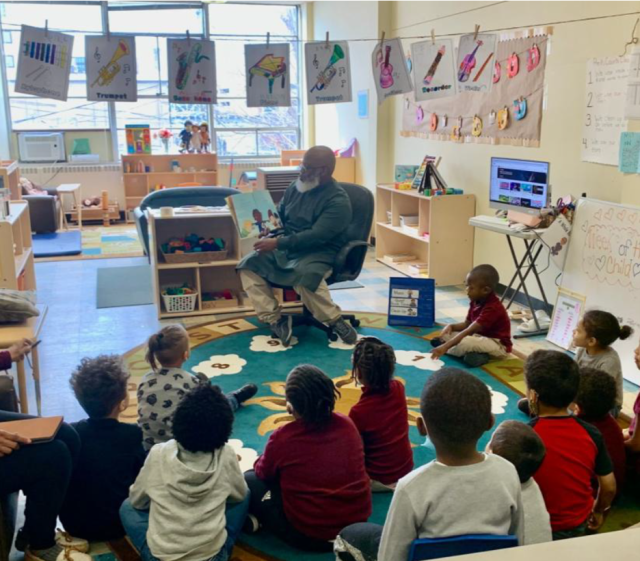 Children sit on the floor, listening to a man read an illustrated story.
