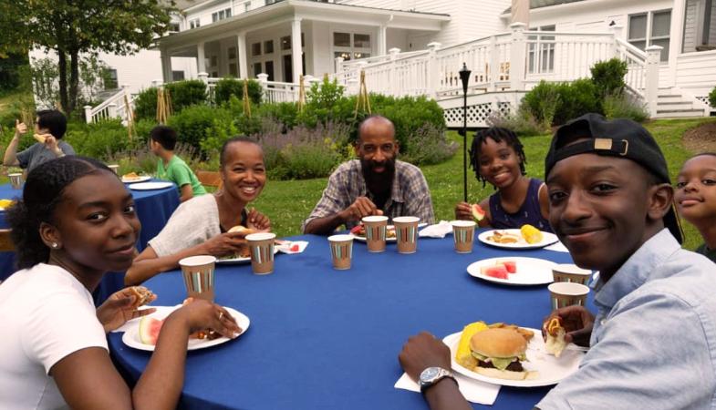 A family enjoys a Fourth of July lunch at round tables in the Retreat Center's yard