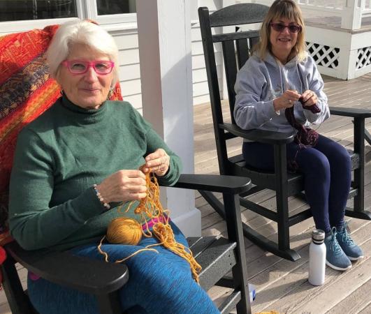 Two women knit in the rocking chairs on the porch