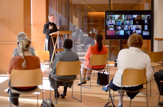 Members of the congregation sit in chairs with WebEx screen and Vicar speaking at Discovery