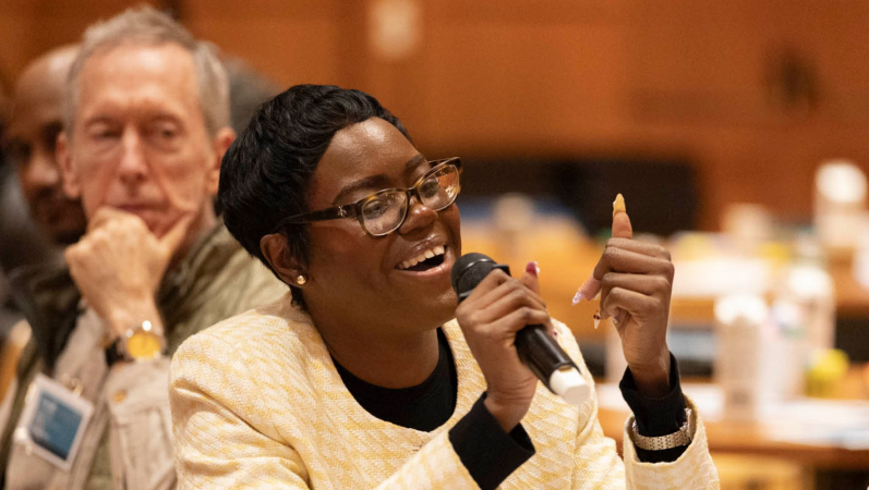Woman speaks into a microphoto while sitting at a table in the audience of the Racial Justice Initiative Grantee Convening