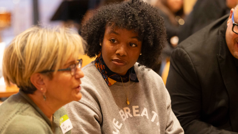 Woman watches another woman talking while they're sitting at a table.
