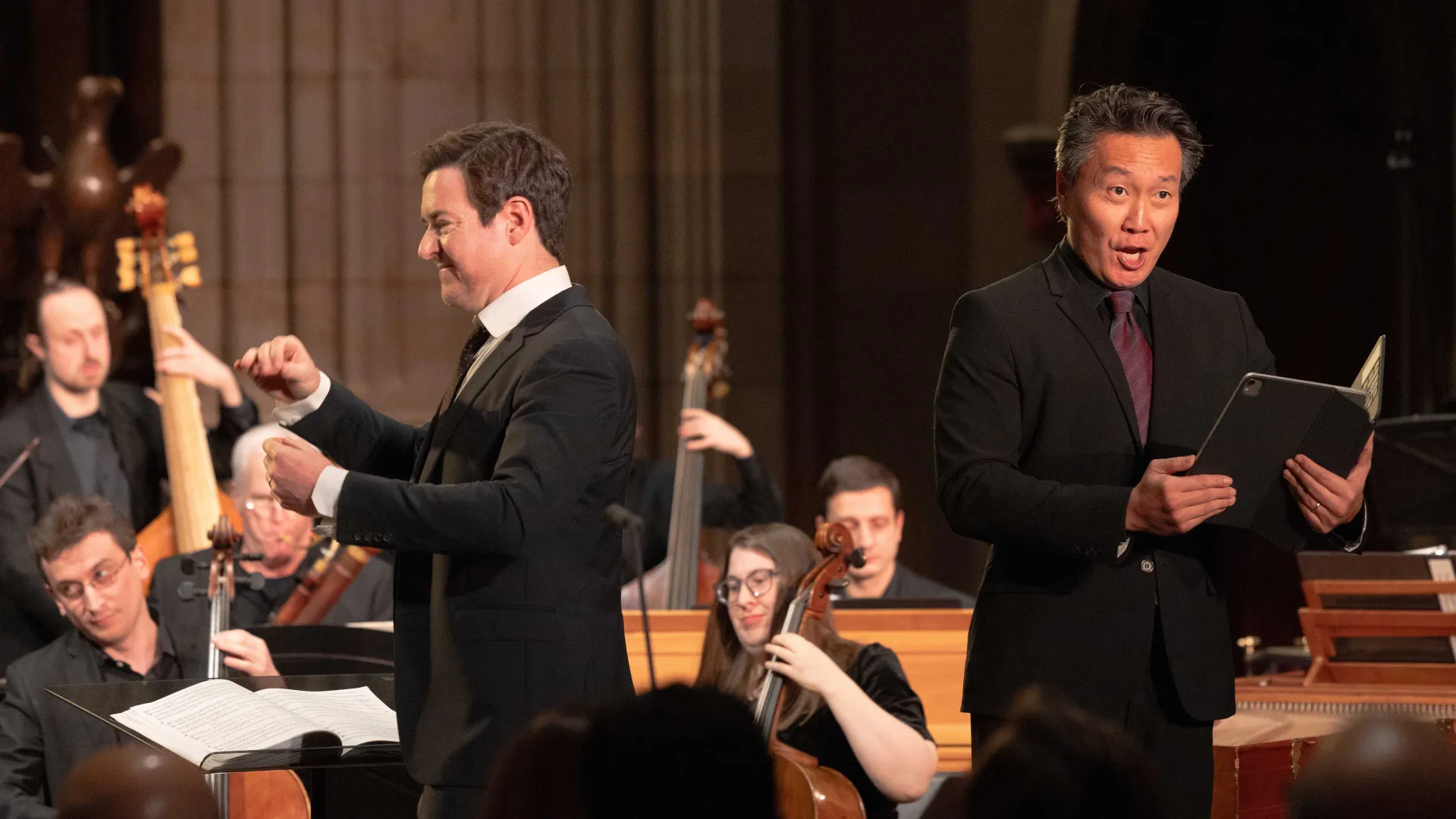 Man on left conducts an orchestra and singers, man on right sings passionately