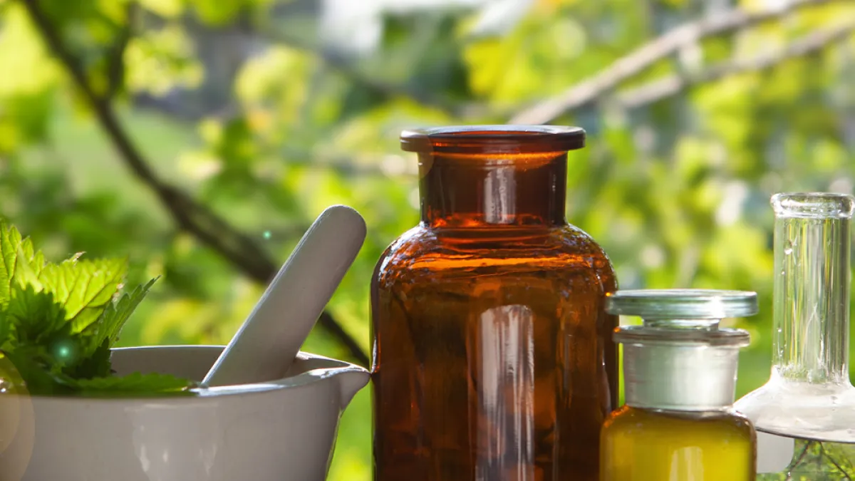 A mortar and pestle filled with leaves and vintage apothecary bottles filled with oils against a backdrop of green trees