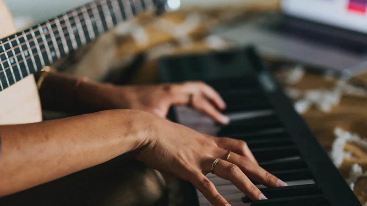 A person holds a guitar and plays a keyboard with a laptop in the background