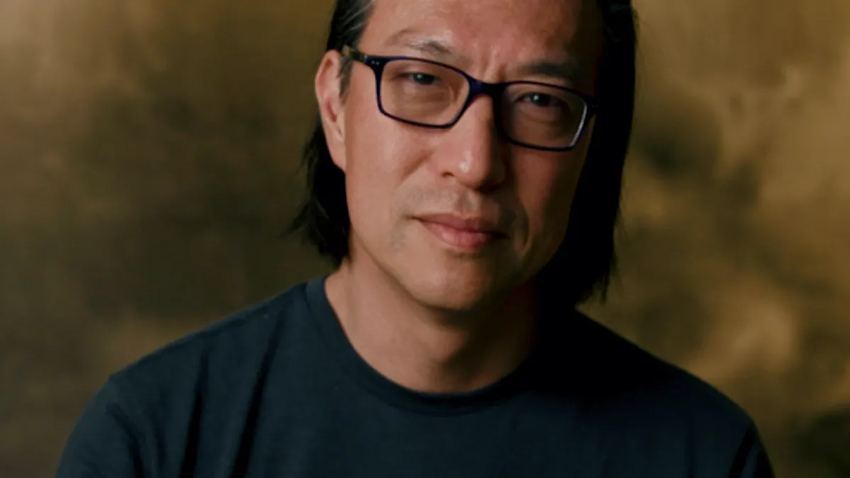 Makoto Fujimura stands in front of an ochre background, wearing glasses and a black crewneck shirt. He smiles slightly with his head tilted to the right.