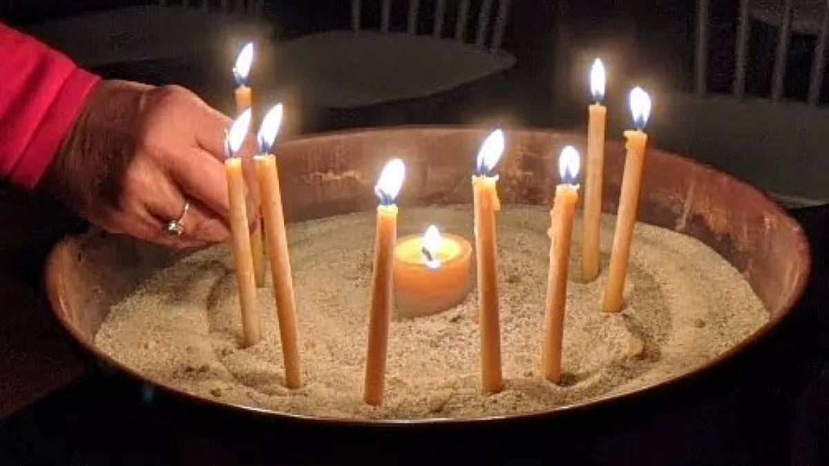 One hand straightens a lit taper in a brass basin filled with sand. A total of 8 tan tapers and a tan tea candle are lit in the basin.