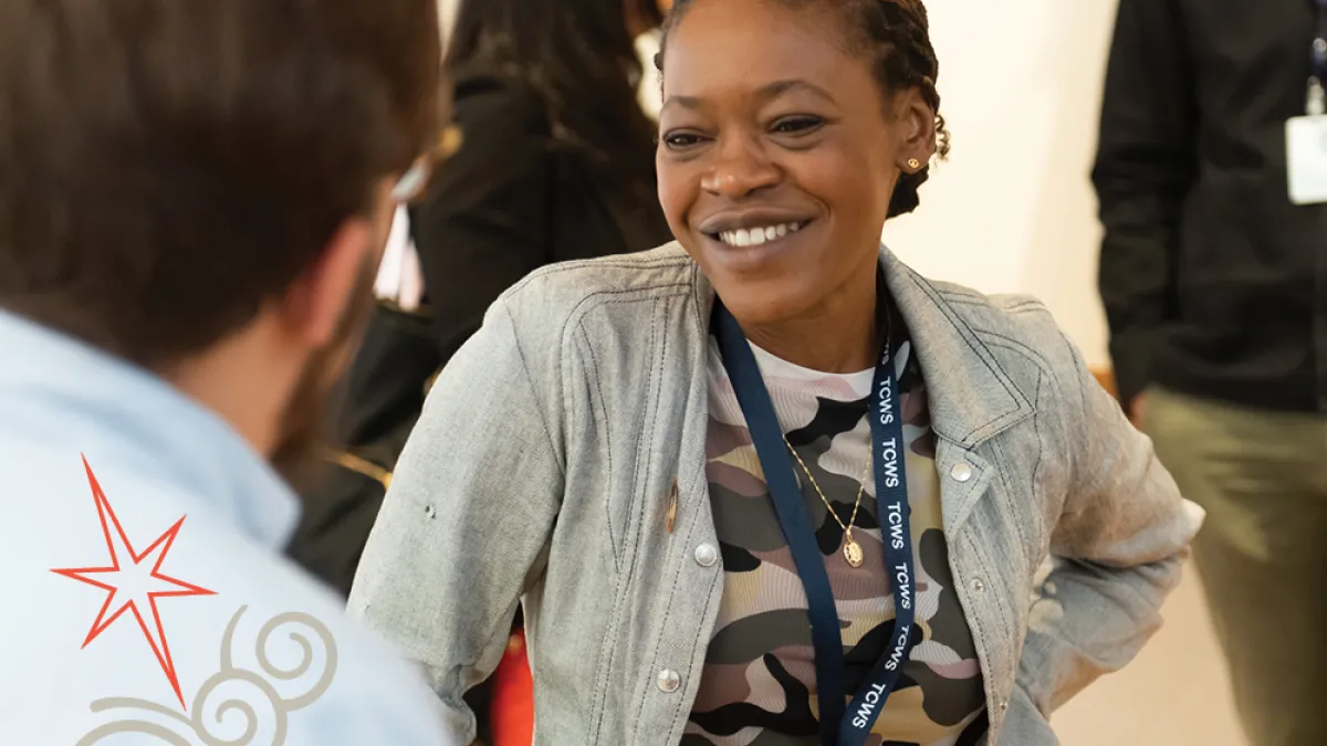Trinity Leadership Fellow Chiseche Mibenge smiles while in conversation with a person whose back is to us. Chiseche is a Black woman wearing a light denim shirt over an abstract colored tee and lanyard on her neck. 