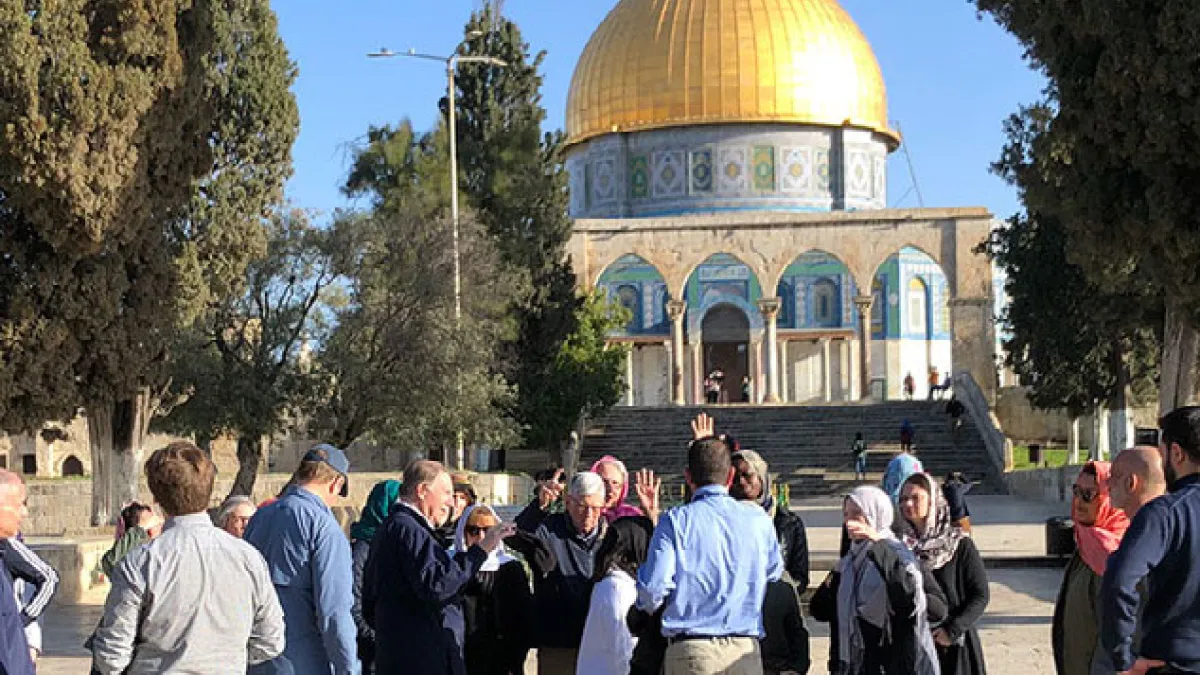 A group of people stand in a plaza under a bright blue, cloudless sky on a sunny day. They are visiting an historic temple in the Holy Land with a golden dome.