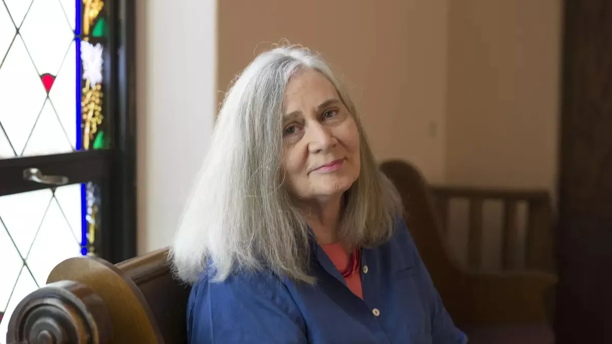 Marilynne Robinson sits on a pew and turns to face the camera, while light from a stained glass window shines on her. She has grey shoulder length straight hair, fair skin, and wears a blue button-down shirt with coral undershirt.