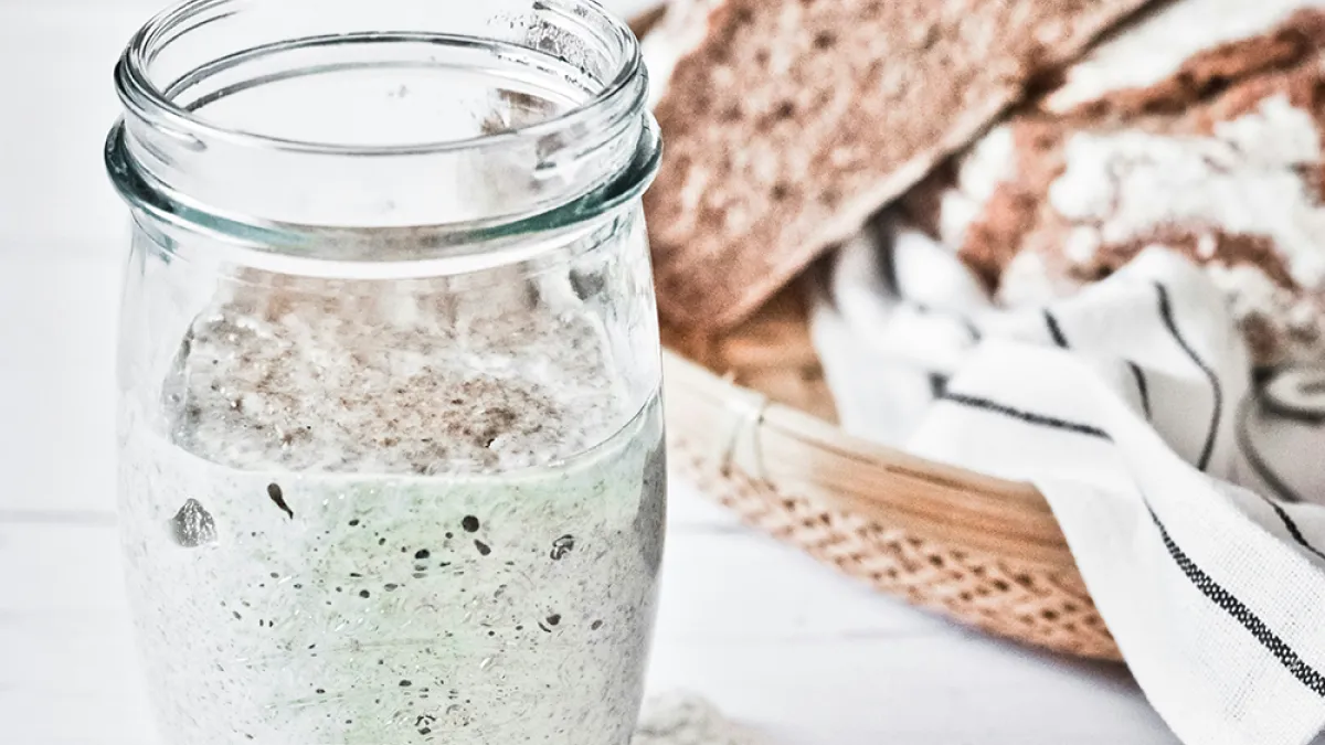 An image of sourdough starter in a glass jar, with a sliced loaf of bread in the background.