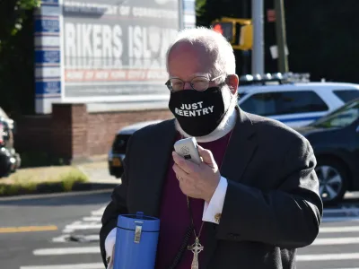 The Rt. Rev. Andrew M.L. Dietsche stands in front of the Rikers Island sign with a megaphone and a mask reading "Just Reentry."