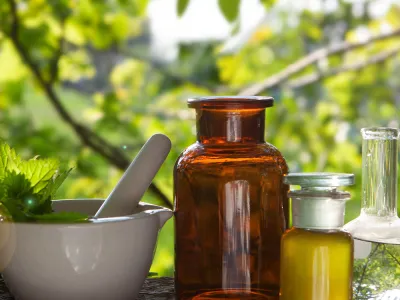 A mortar and pestle filled with leaves and vintage apothecary bottles filled with oils against a backdrop of green trees
