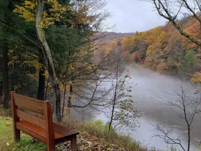 A wooden bench on a hill overlooking a foggy river in the middle of a forest of autumnal foliage