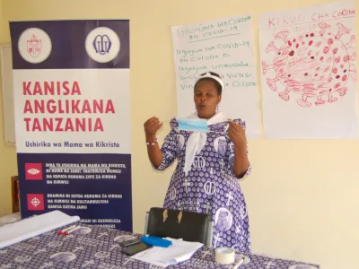 Margareth Massawa demonstrates proper face mask use, holding a mask in her hand while standing in front of informational signs about COVID-19 transmission.