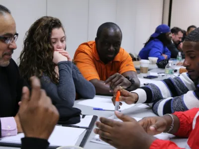 A group of young Black men and a White woman are gathered in a circle in cheerful discussion. Another group of men can be seen in the background. 