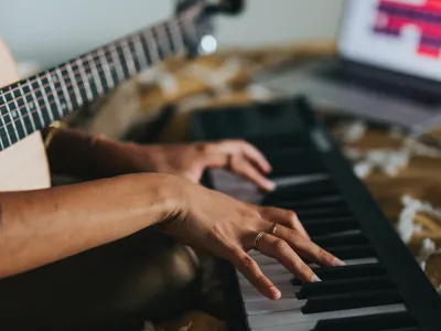 A person holds a guitar and plays a keyboard with a laptop in the background