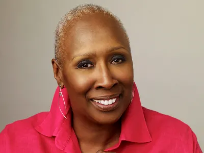 Image of a woman smiling in front of a beige background. She has closely cropped hair, and is wearing a hot pink linen shirt with a popped collar. 