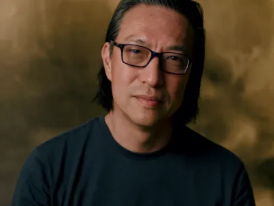 Makoto Fujimura stands in front of an ochre background, wearing glasses and a black crewneck shirt. He smiles slightly with his head tilted to the right.