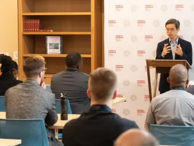 The Rev. Tony Tian-Ren Lin, PhD speaks to members of the inaugural cohort of Trinity Leadership Fellows as they sit in the audience. He stands in front of a step-and-repeat that features Trinity Church Wall Street branding on it.