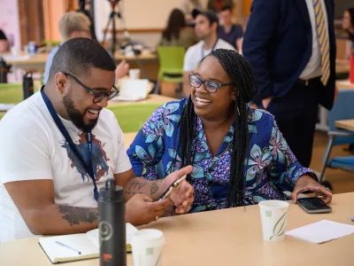 Two people are laughing together during paired conversation. The Black man wears a white tee with a graphic print, and glasses, while the Black woman has braided hair and wears a blue floral dress and glasses.