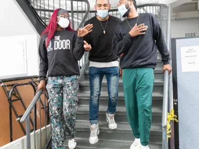 Masked young people are laughing in conversation while walking down a staircase. The young Black woman at left has pink hair and wears glasses and a sweatshirt that reads "The Door." The two young Black men wear casual clothes (jeans, sneakers, and sweatshirts). All three wear white sneakers.