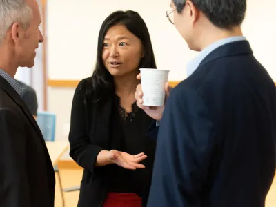 An Asian woman is in conversation with a White man and Asian Man. She has dark hair and is wearing a black cardigan, black lace-lined blouse, and red skirt. The White man has white hair and is wearing a dark grey suit. The Asian man wears glasses, a navy suit, and is holding a white coffee cup.