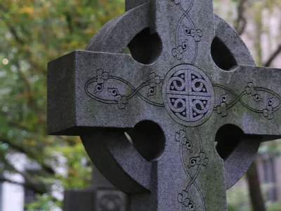 A close-up photograph of a cross-shaped headstone in Trinity Churchyard with trees and Trinity Church in the background