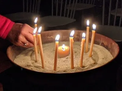 One hand straightens a lit taper in a brass basin filled with sand. A total of 8 tan tapers and a tan tea candle are lit in the basin.