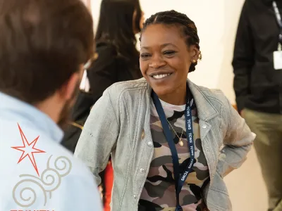 Trinity Leadership Fellow Chiseche Mibenge smiles while in conversation with a person whose back is to us. Chiseche is a Black woman wearing a light denim shirt over an abstract colored tee and lanyard on her neck. 