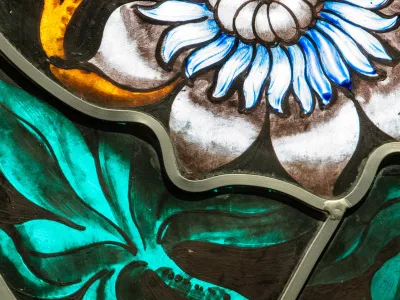 A close-up photo of stained glass depicting a white and blue flower with vivid green leaves