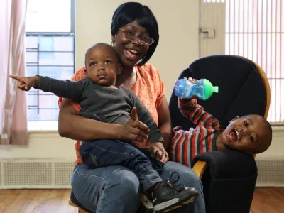 A Black mother wearing glasses and her hair in a black bob smiles with her two children. One child smiles and leans over with his sippy cup in hand, while the other sits in her lap with a serious expression and points to the left.