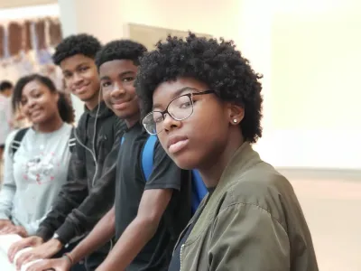 Four black children stand in a row facing the camera. The three children in the background smile softly, while the child in the foreground wears glasses and looks seriously at the camera.