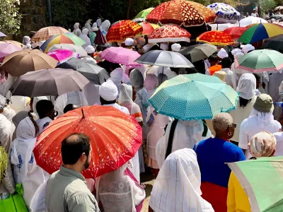 A crowd of people stand on a path. Many hold colorful umbrellas or wear scarves to protect themselves from the sun.