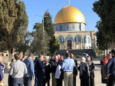 A group of people stand in a plaza under a bright blue, cloudless sky on a sunny day. They are visiting an historic temple in the Holy Land with a golden dome.