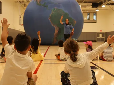Children in the Trinity Commons gym raise their hands in front of an inflatable globe as part of a science lesson during the 2023 Children's Summer Program