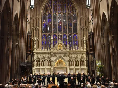 The Choir of Trinity Wall Street performs in Trinity Church with stained glass window behind them.