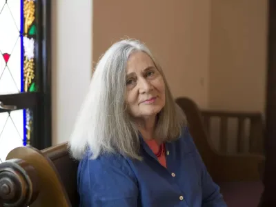 Marilynne Robinson sits on a pew and turns to face the camera, while light from a stained glass window shines on her. She has grey shoulder length straight hair, fair skin, and wears a blue button-down shirt with coral undershirt.