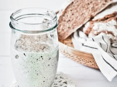 An image of sourdough starter in a glass jar, with a sliced loaf of bread in the background.