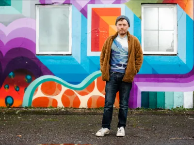 A man in a knit cap, brown cardigan and jeans stands in front of a colorful mural.