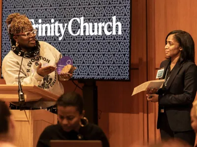 Woman on the left stands behind a podium, holding an award, while speaking to the woman on the right who will soon receive the award.. 