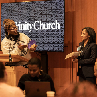 Woman on the left stands behind a podium, holding an award, while speaking to the woman on the right who will soon receive the award.. 
