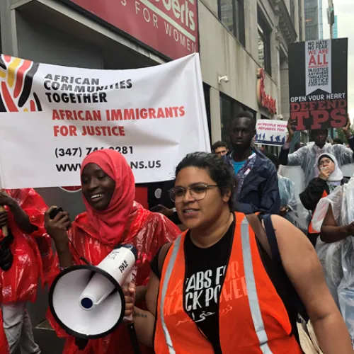 Protestors march on the street. In the foreground, a hijabi holds a megaphone while smiling. Another woman in glasses and an orange vest stands next to her.