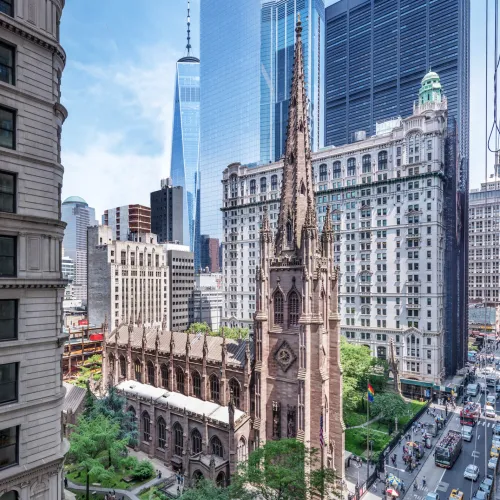 An exterior view of Trinity Church and its Churchyard, surrounded by Trinity Commons, the buildings of downtown Manhattan and Broadway. 