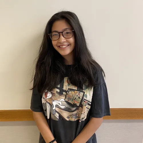 Neyu Higuchi-Crowell faces the screen with a smile. She is wearing glasses and a dark graphic tee.