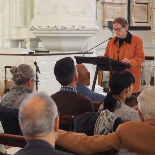 Man in an orange jacket with short dark hair and glasses reads poetry from a lectern in front of an audience at St. Paul's Chapel.