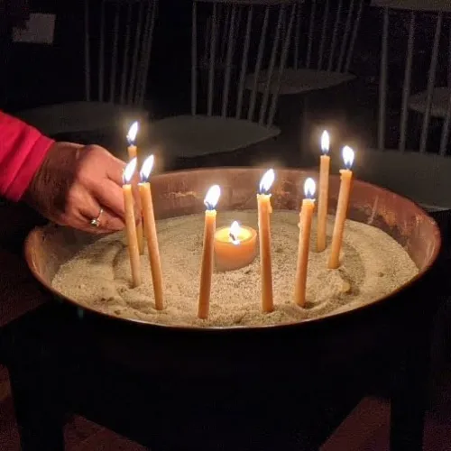 One hand straightens a lit taper in a brass basin filled with sand. A total of 8 tan tapers and a tan tea candle are lit in the basin.