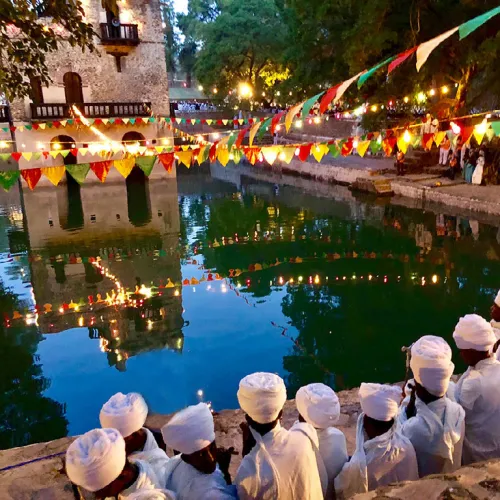 Men in white shirts and turbans face a pool. Streamers with triangular flags criss-cross the water above. There is a stone structure in the background, and the surrounding trees are reflected in the water.