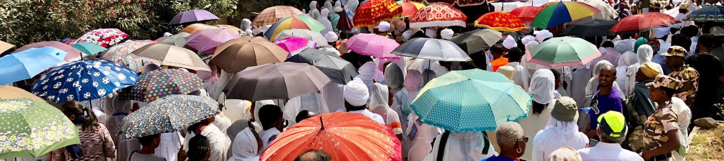 A crowd of people stand on a path. Many hold colorful umbrellas or wear scarves to protect themselves from the sun.